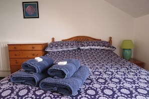 Een net opgemaakt bed met een bedekt met een patroon, kussens, een houten nachtkastje met een groene lamp en een houten dressoir met een foto aan de muur.