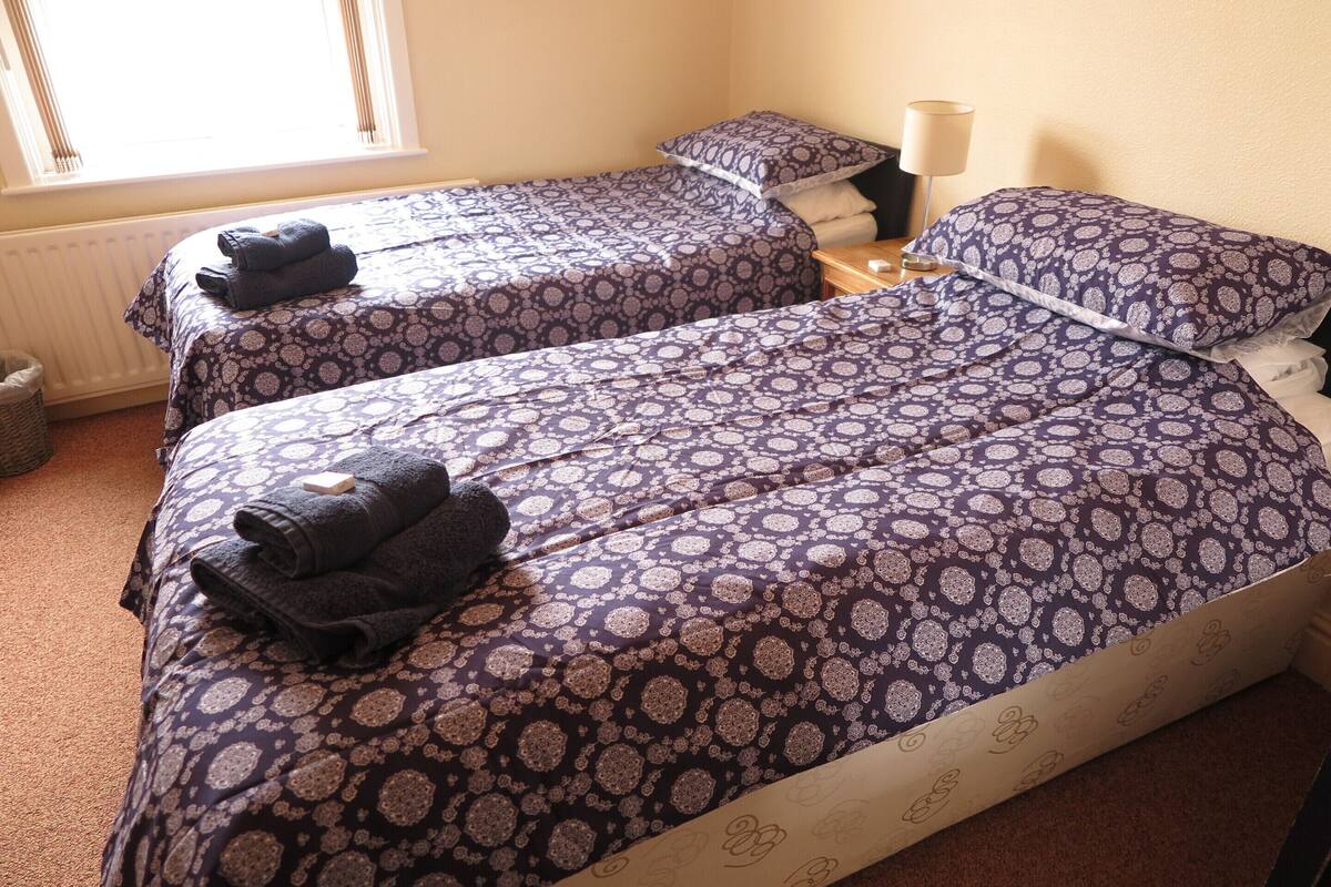 Two twin beds with matching patterned bedding, a bedside table with a lamp, and a window with blinds.