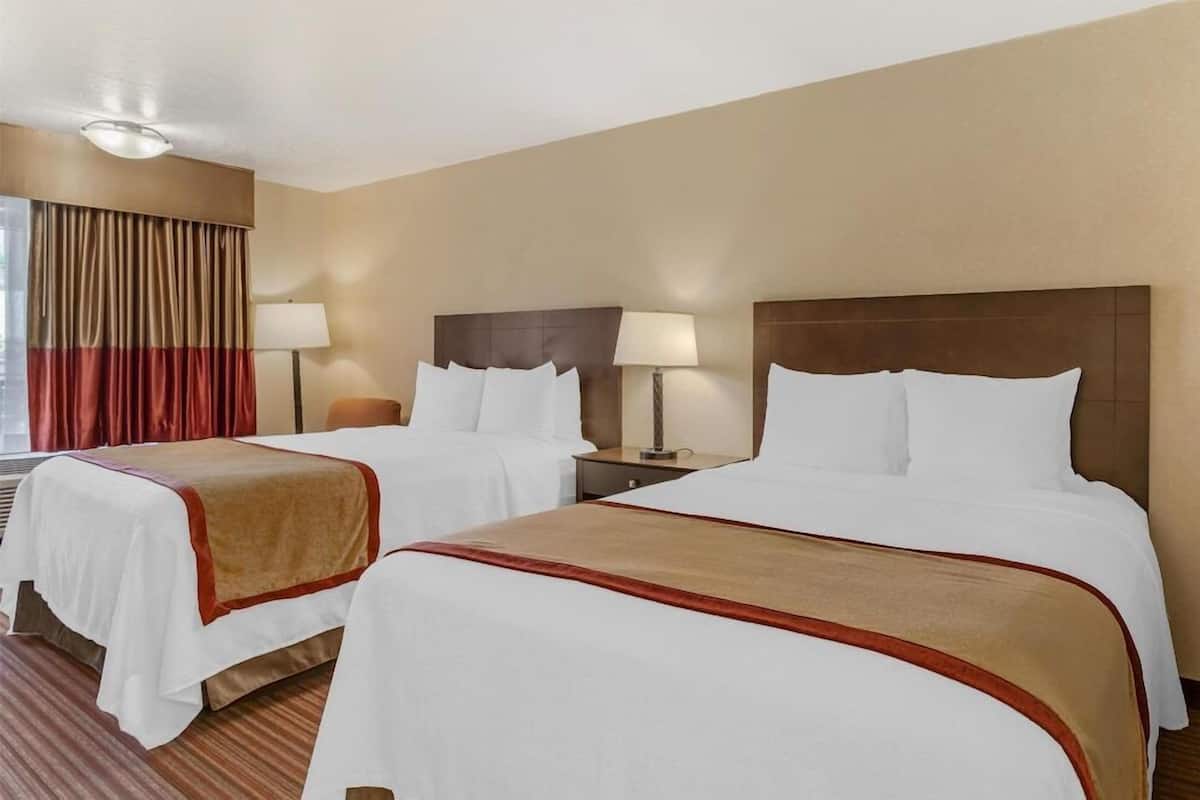 A hotel room with two beds, each with a headboard, bedside tables, lamps, and a window with curtains.
