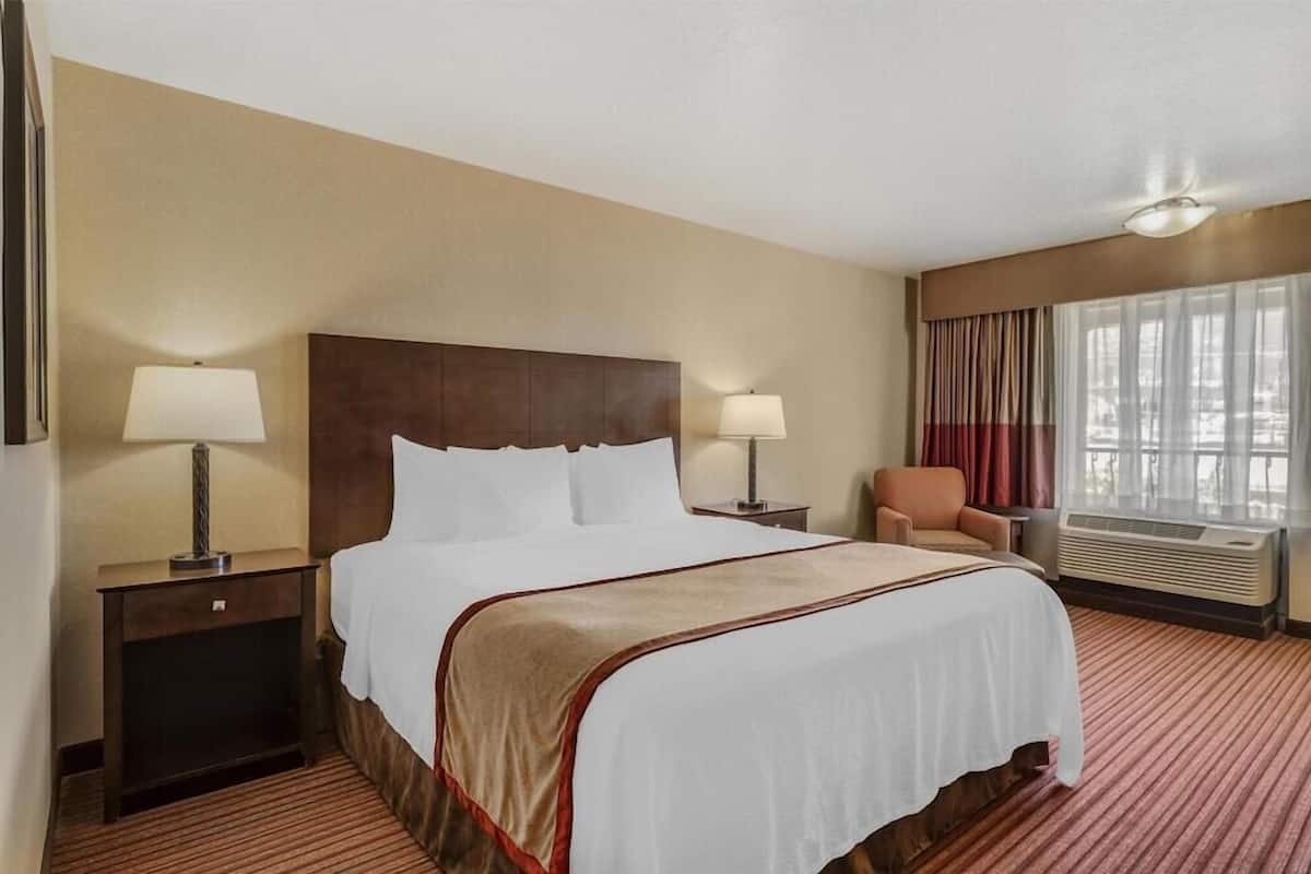 A hotel room with a large bed, two bedside tables with lamps, a chair, a window with curtains, and an air conditioning unit.