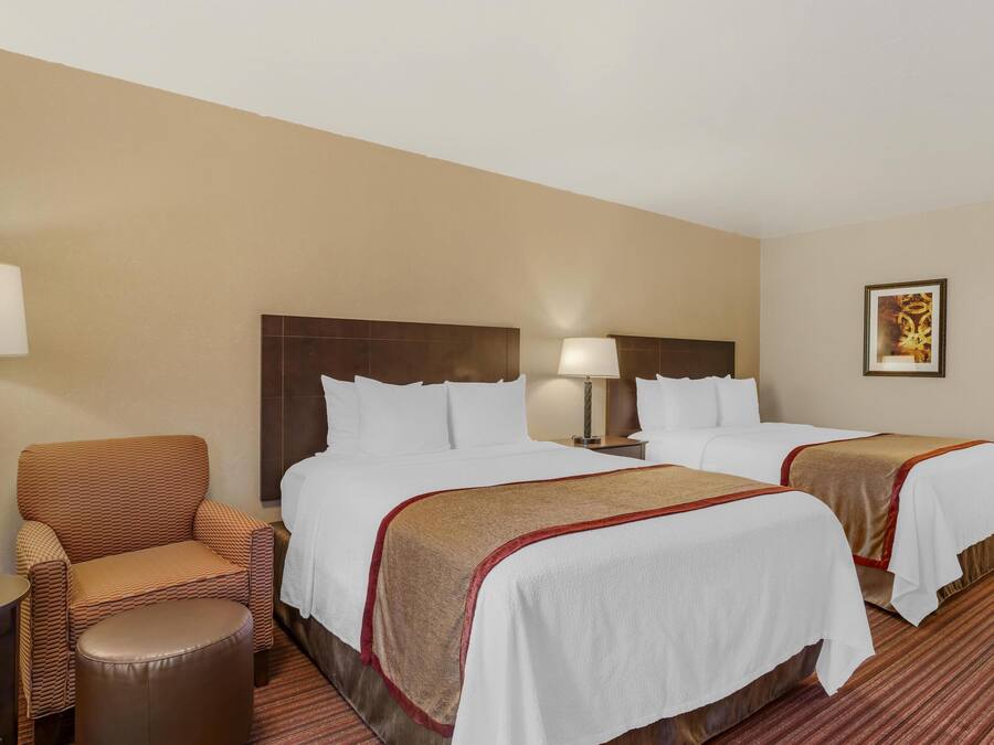 A hotel room with two beds, a chair, a small table, and a lamp.