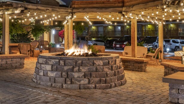 Ein Außenbereich mit einer Steinkaminanlage, einer Holzpergola, Lichterketten und Sitzgelegenheiten.