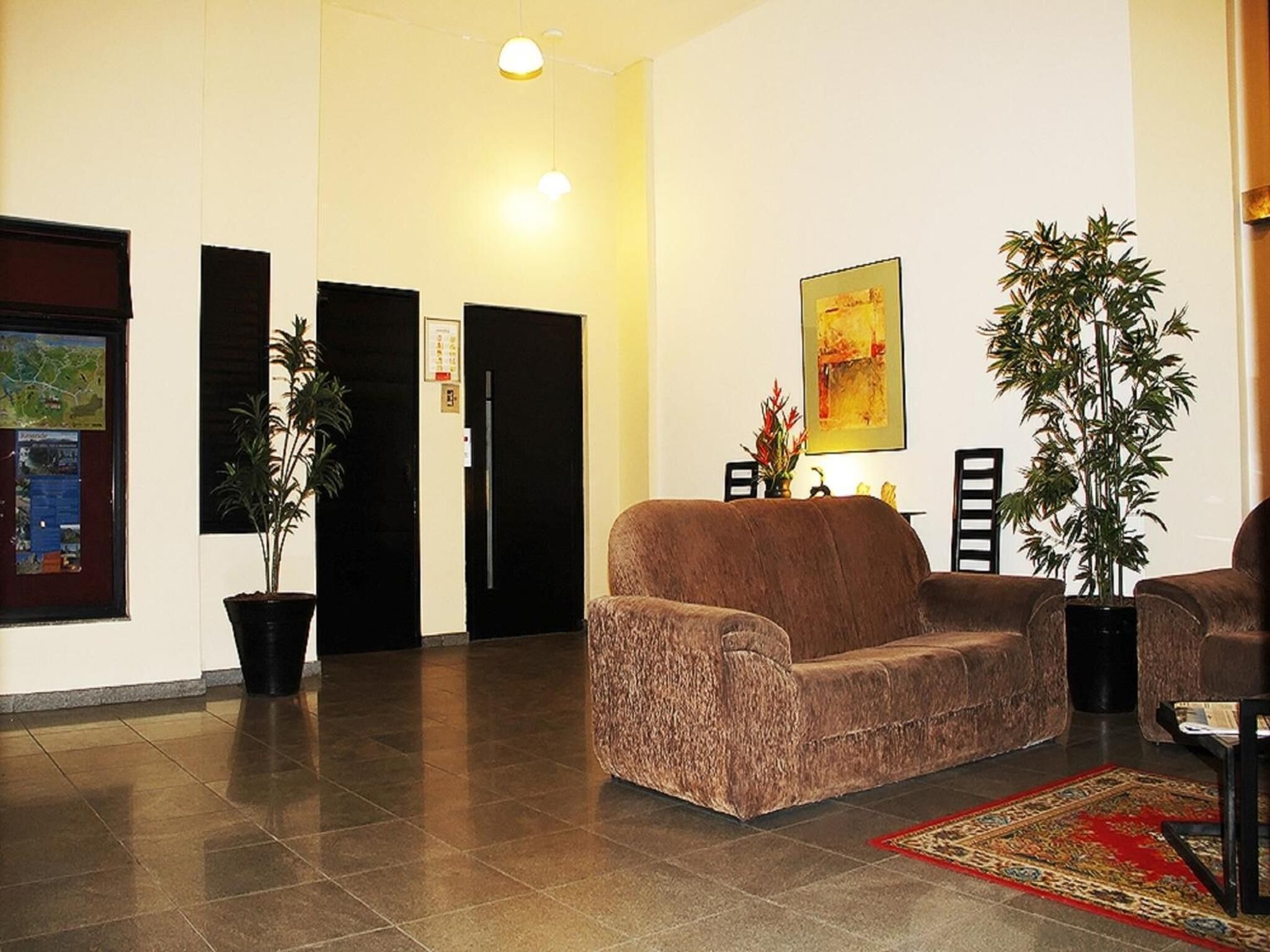 A lobby with a brown sofa, two potted plants, a red rug, and a framed picture on the wall.