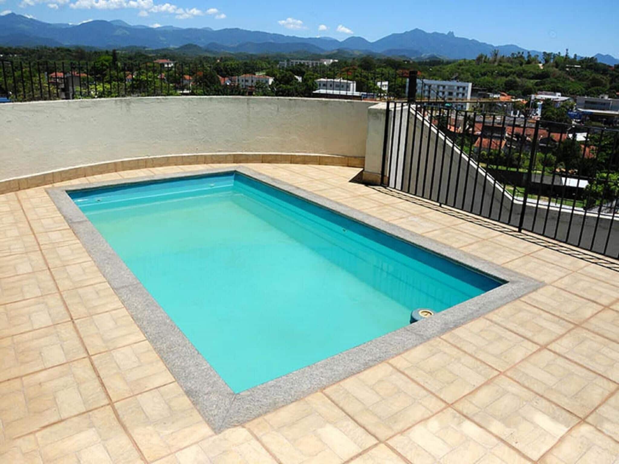 Une piscine rectangulaire, bordée d’une terrasse carrelée et entourée d’une clôture en métal noir, avec en arrière-plan un paysage de montagnes et d’immeubles d’habitation.