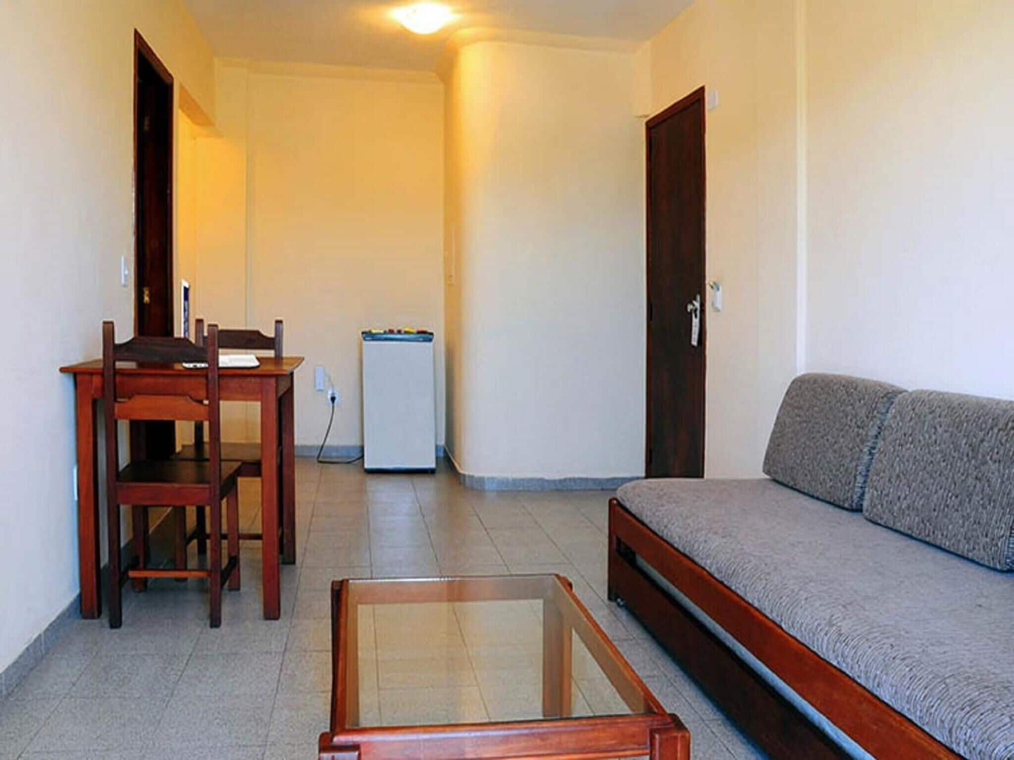 A compact living space with a wooden dining table, a glass-top coffee table, a grey sofa, and a small white refrigerator.