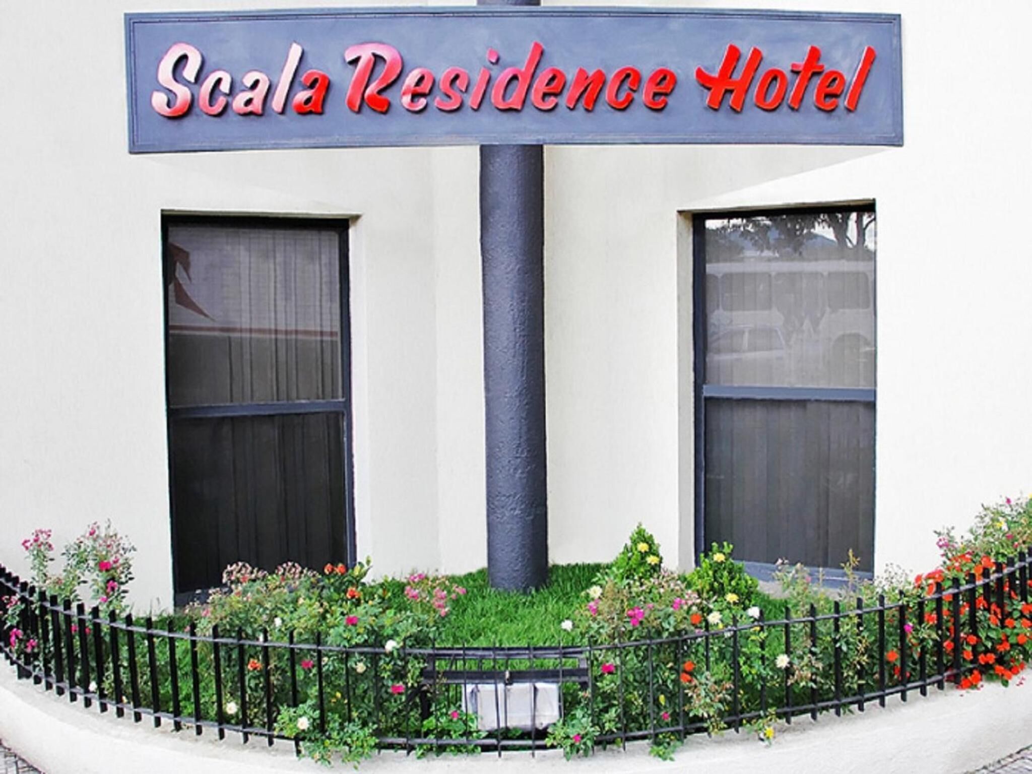 Señal de hotel que indica 'Scala Residence Hotel', con dos ventanas y un jardín de flores al frente.