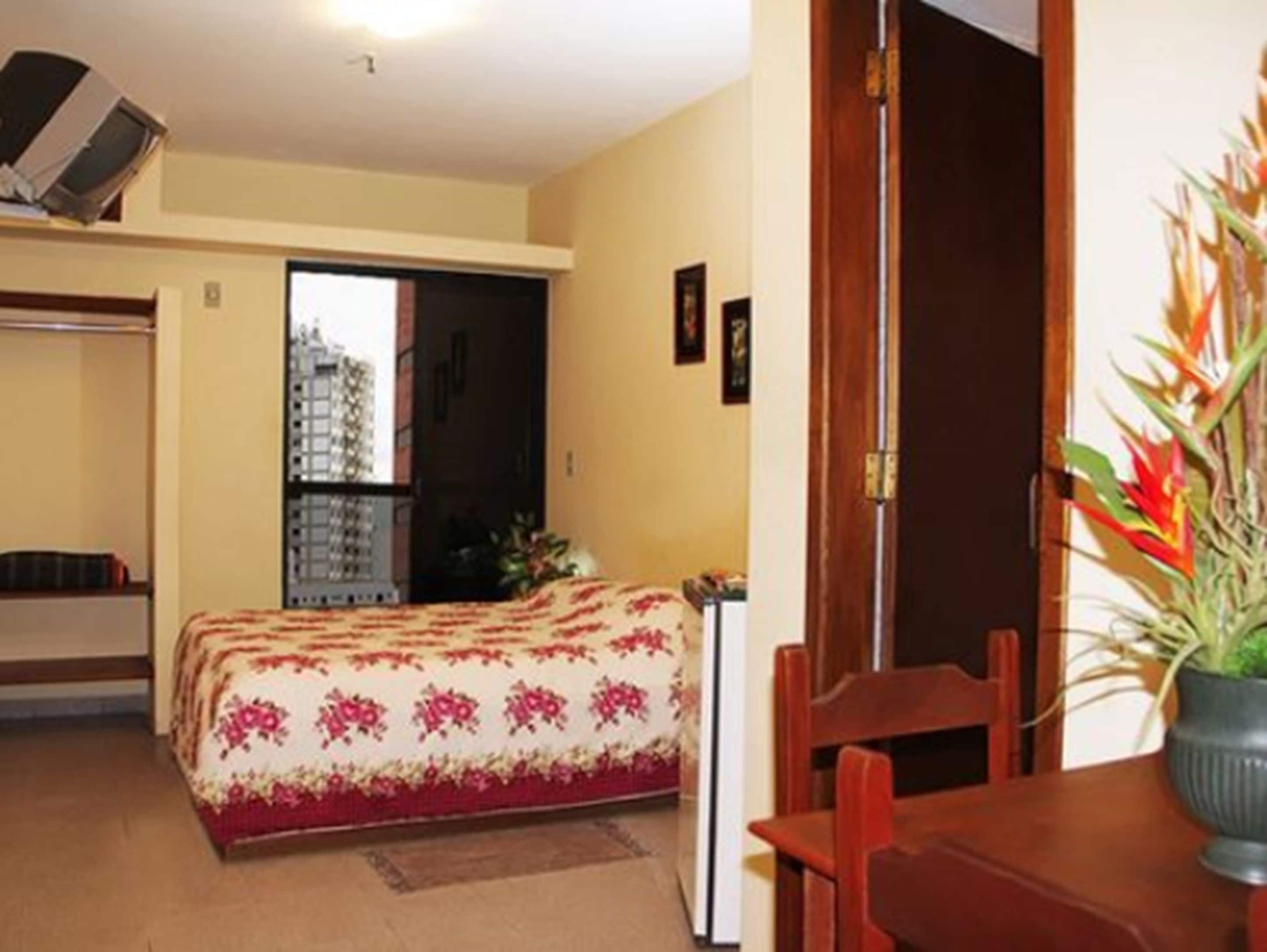 A small, cozy room with a bed, a wooden chair, a table, and a potted plant.