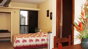 A small, cozy room with a bed, a wooden chair, a table, and a potted plant.