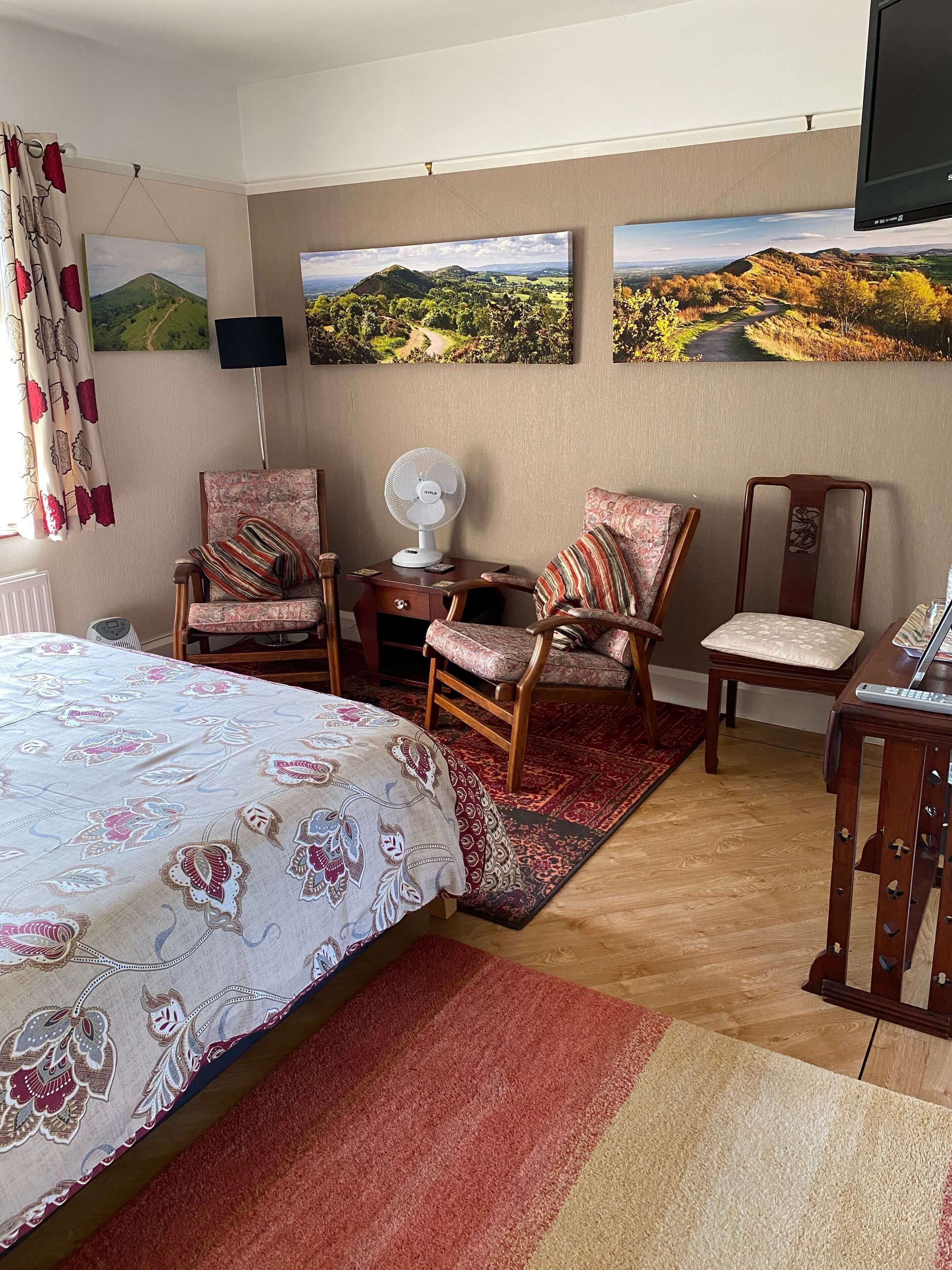 A bedroom with a bed, two chairs, a desk, a fan, and three landscape paintings on the wall.