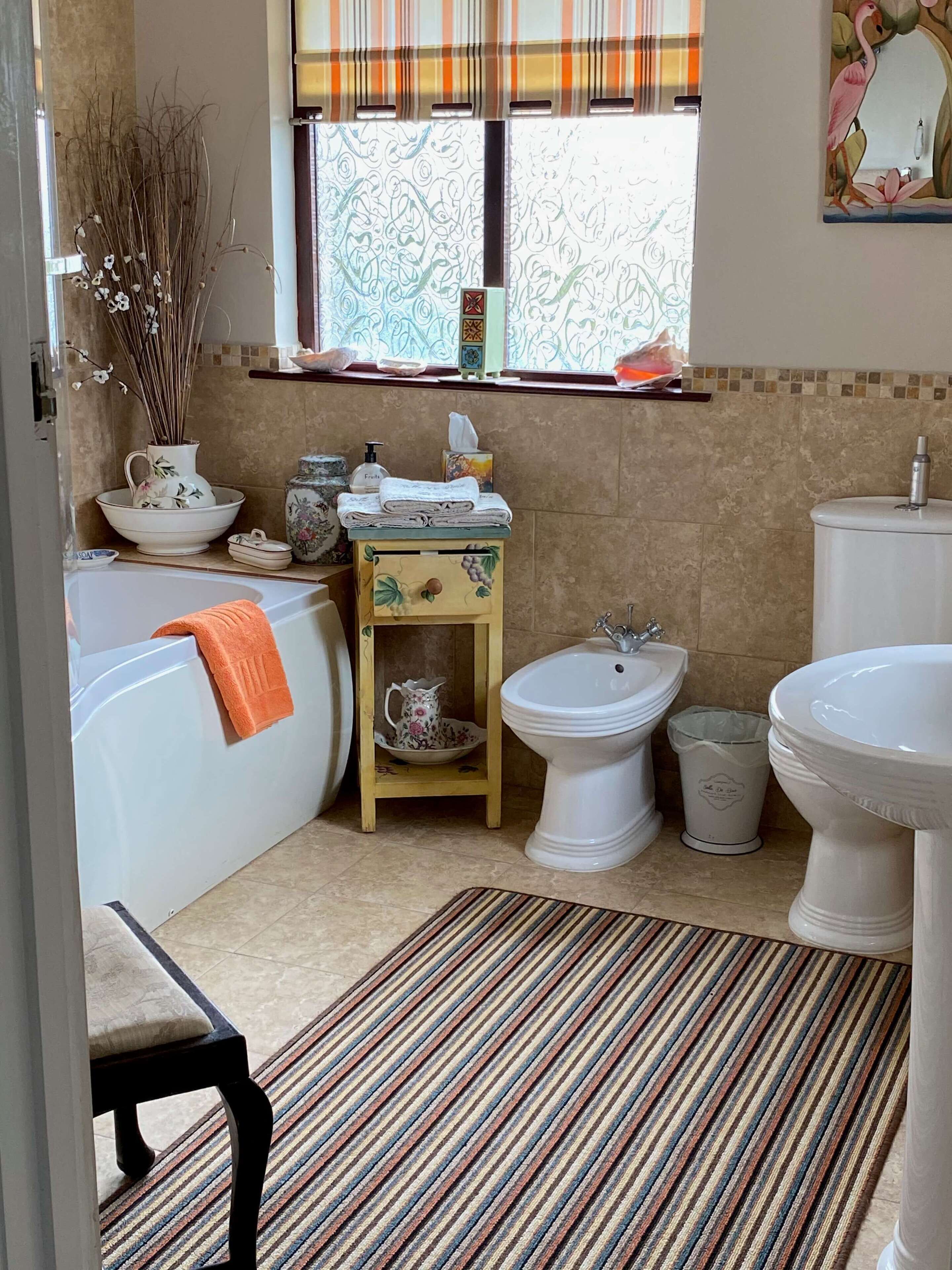 A bathroom with a bathtub, bidet, sink, and a window with a view of trees. There is a striped rug on the floor and a small shelf with toiletries.