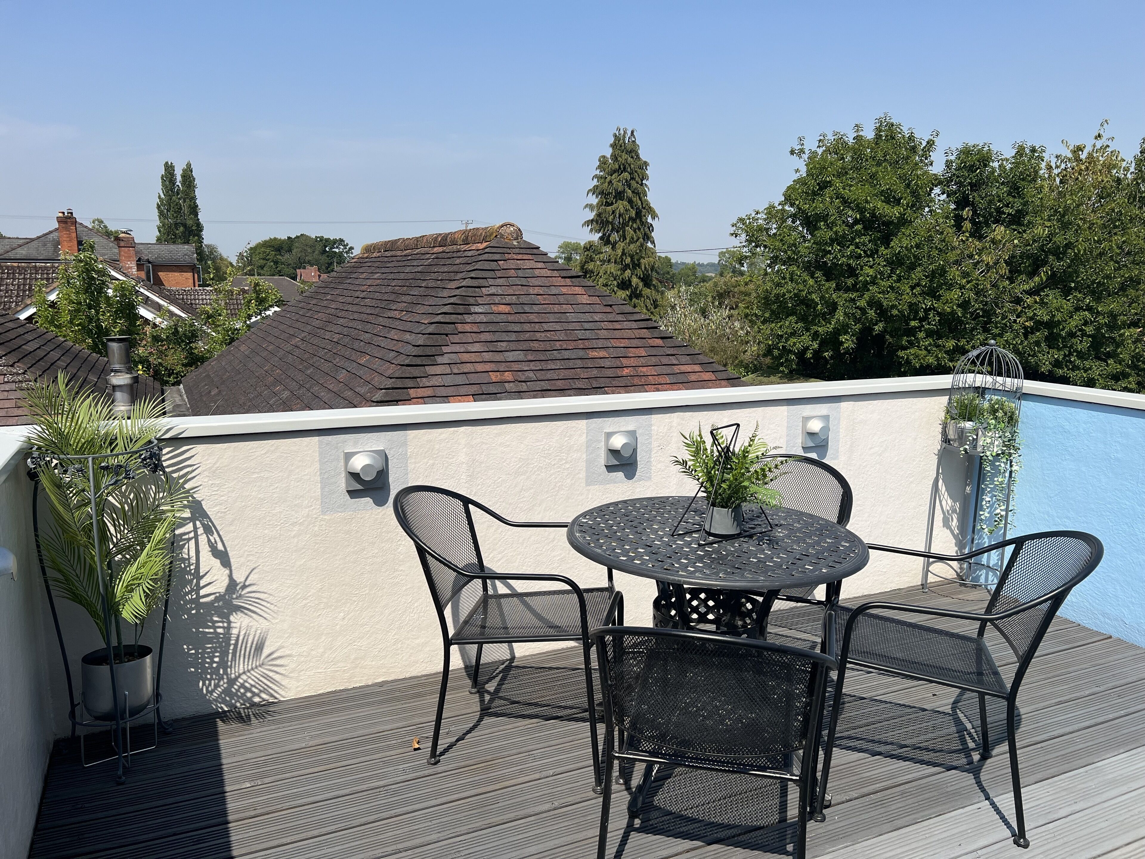 Eine Dachterrasse mit einem runden Tisch und vier Stühlen, umgeben von Pflanzen in Töpfen und Blick auf benachbarte Gebäude und Bäume.