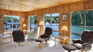 En træhytte med store vinduer med udsigt over en sø, møbleret med læderstole og et lille bord.