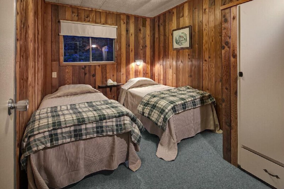 Una habitación con paneles de madera, dos camas con ropa de cama escocesa, una luz nocturna, una ventana con vistas y una puerta.
