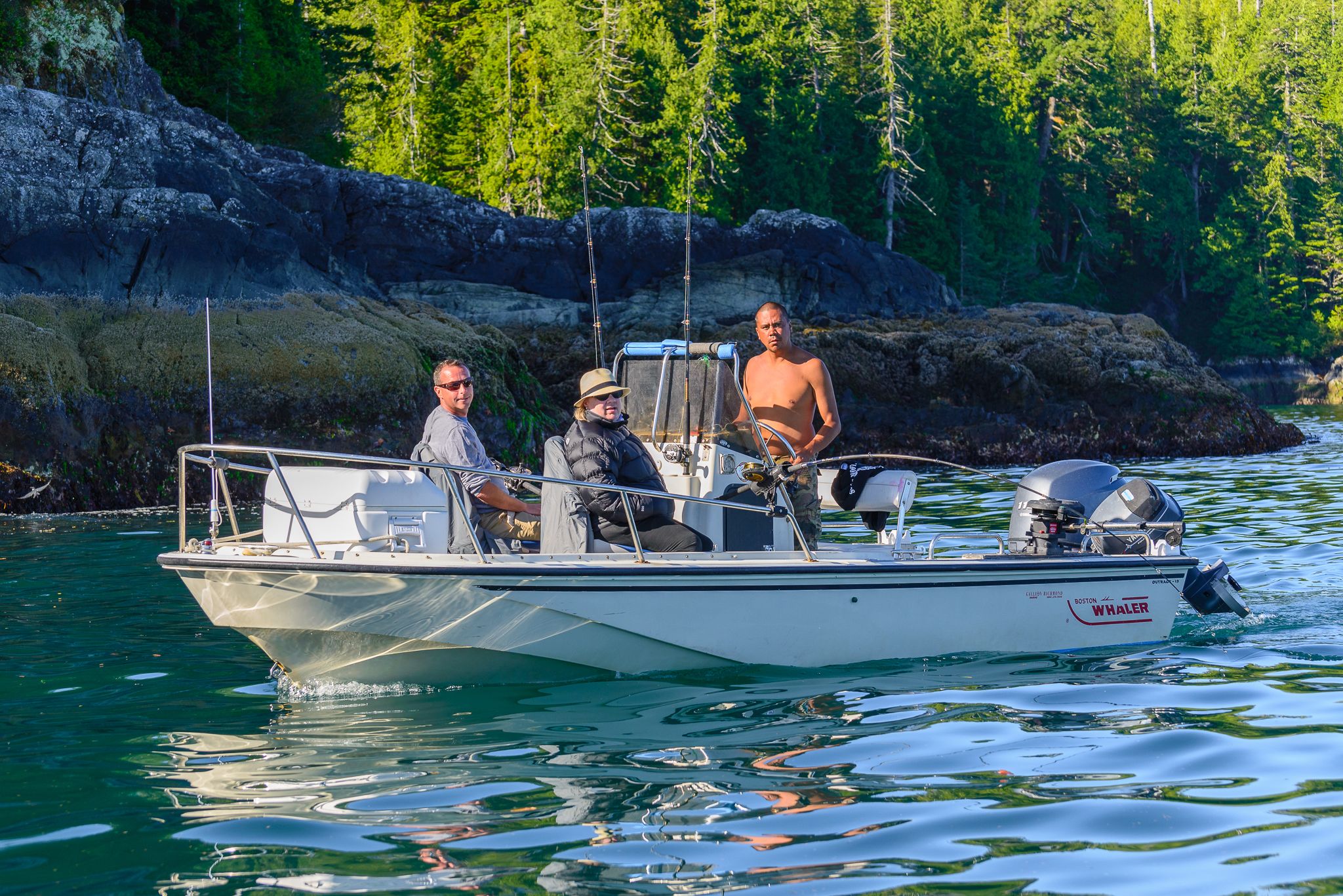 A boat with three people on board, a motor, and a rocky shoreline with trees in the background.