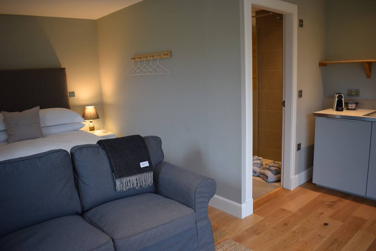 A bedroom with a bed, a grey sofa, a bedside table with a lamp, a wooden coat rack, and a bathroom visible through an open door.