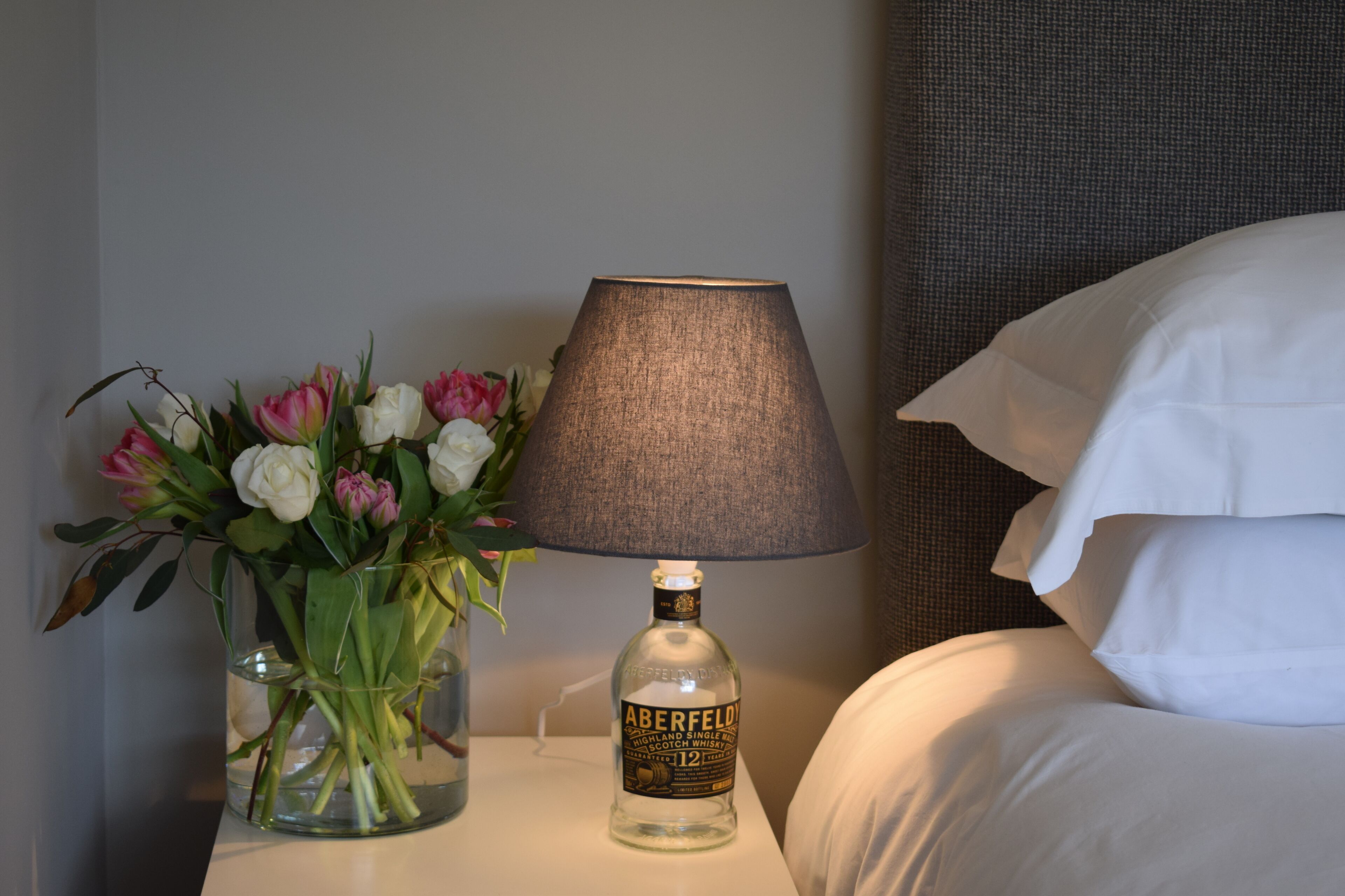 A hotel room with a bedside table featuring a bottle of Aberfeldy whisky, a vase with flowers, and a lamp.
