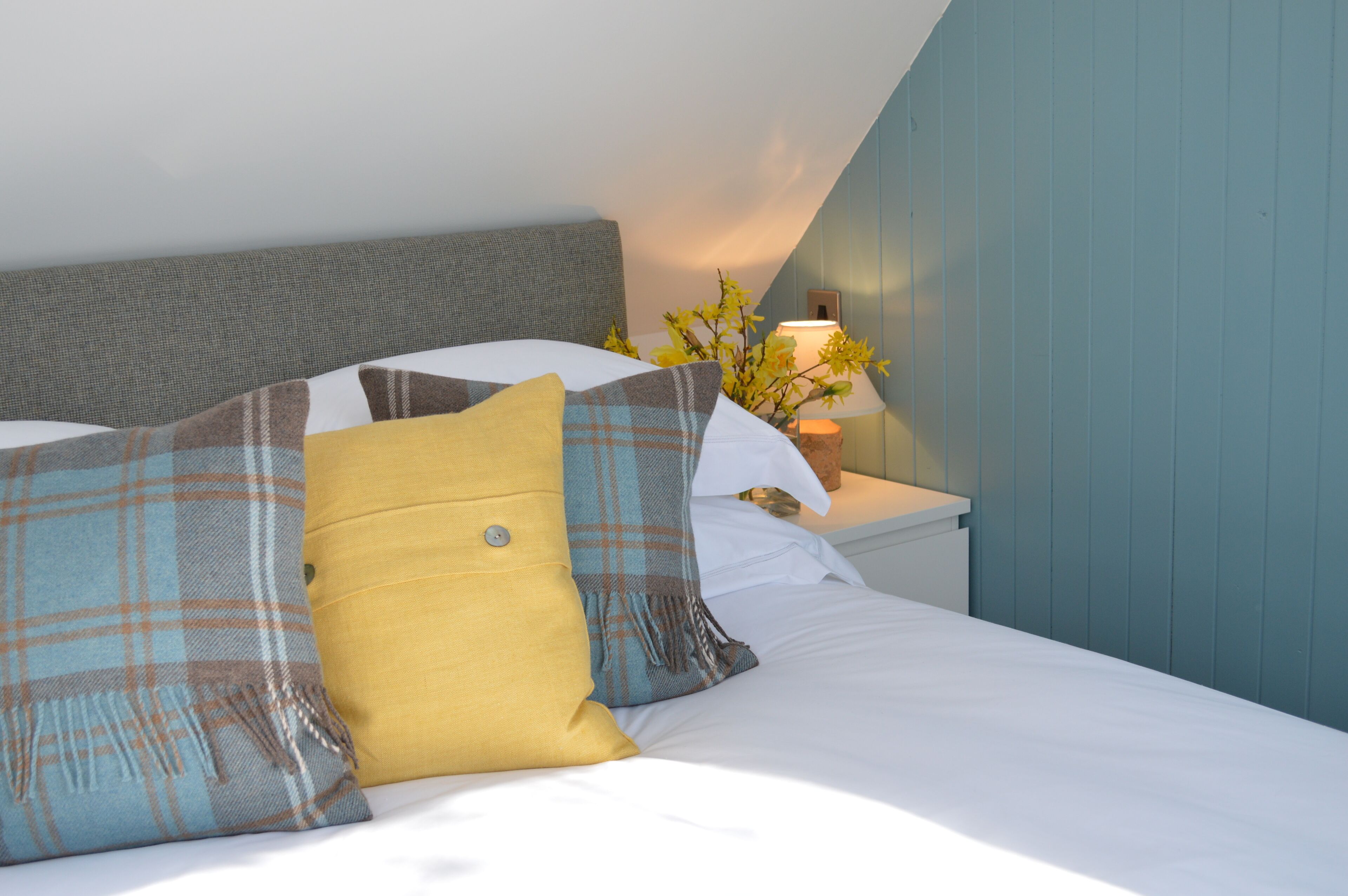A neatly made bed with plaid and solid-colored pillows, a bedside lamp, and a vase with yellow flowers.