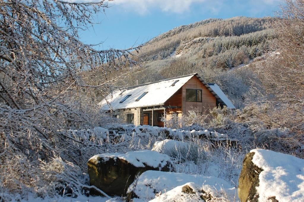 Une cabane au toit en pente, entourée d’arbres couverts de neige, avec une chaîne de montagnes en arrière-plan.