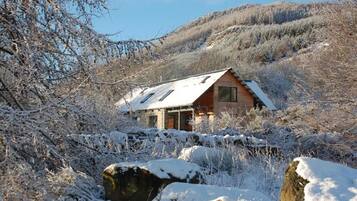 Une cabane au toit en pente, entourée d’arbres couverts de neige, avec une chaîne de montagnes en arrière-plan.
