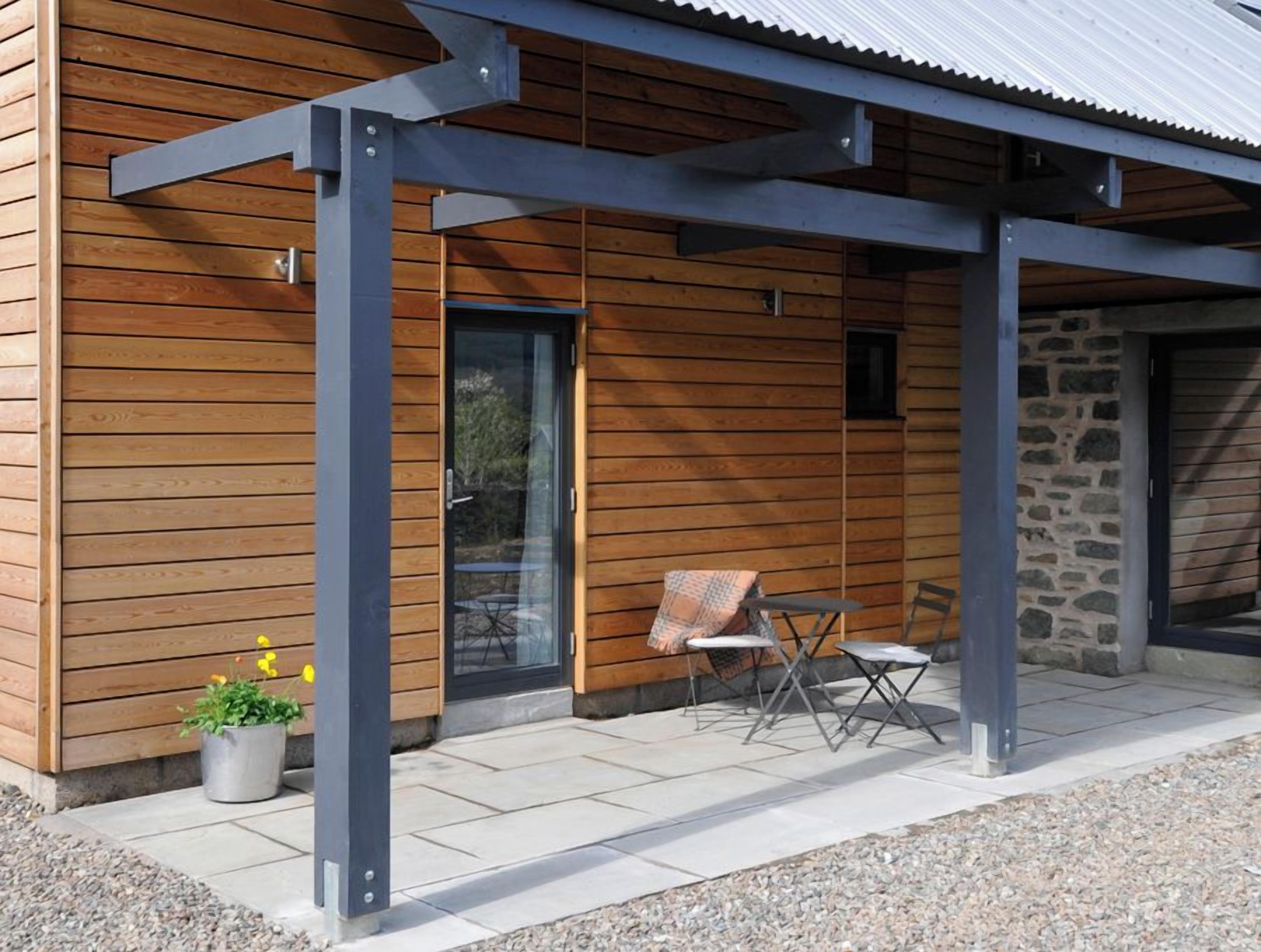 En moderne bygning med træfacade, en stenvæg og en overdækket terrasse med to klapstole og et bord.