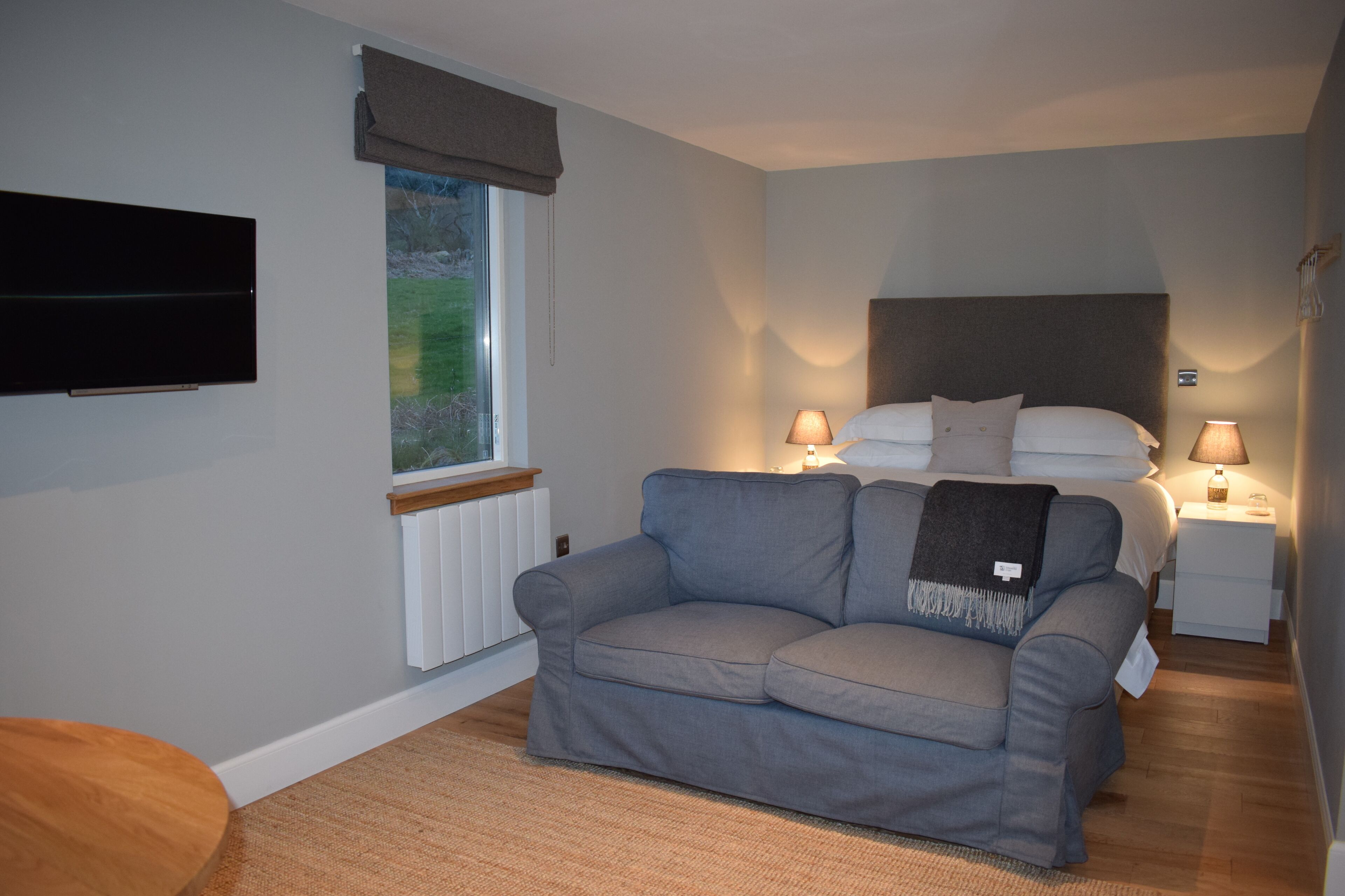 Un dormitorio moderno con un sofá gris, una cama con ropa blanca, una mesita de noche con una lámpara y un televisor montado en la pared.