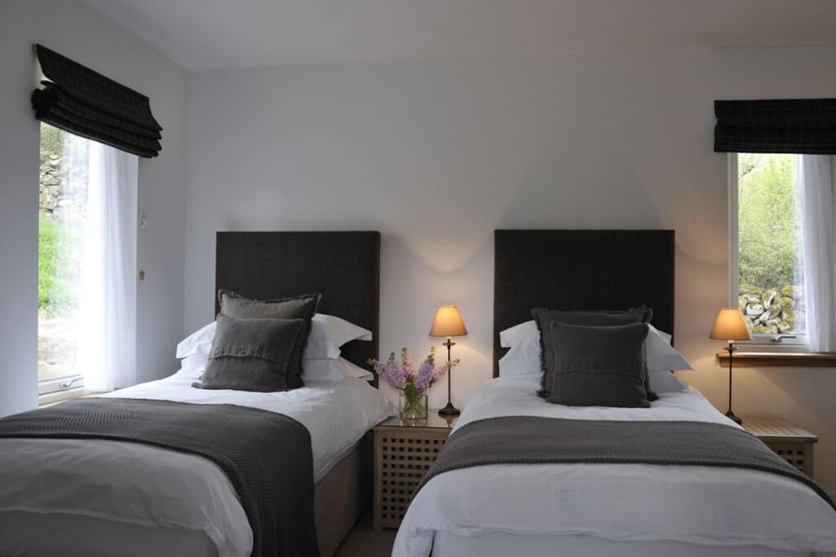 A hotel room with two beds, each with a dark headboard, white bedding, and a small table between them with a lamp and a vase of flowers.