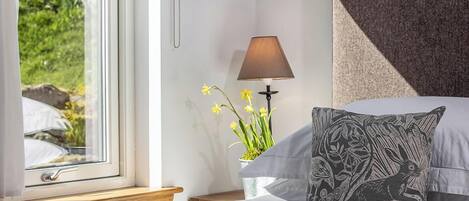 A bedroom with a bed, a bedside table with a lamp, a window with a view of greenery, and a decorative pillow.