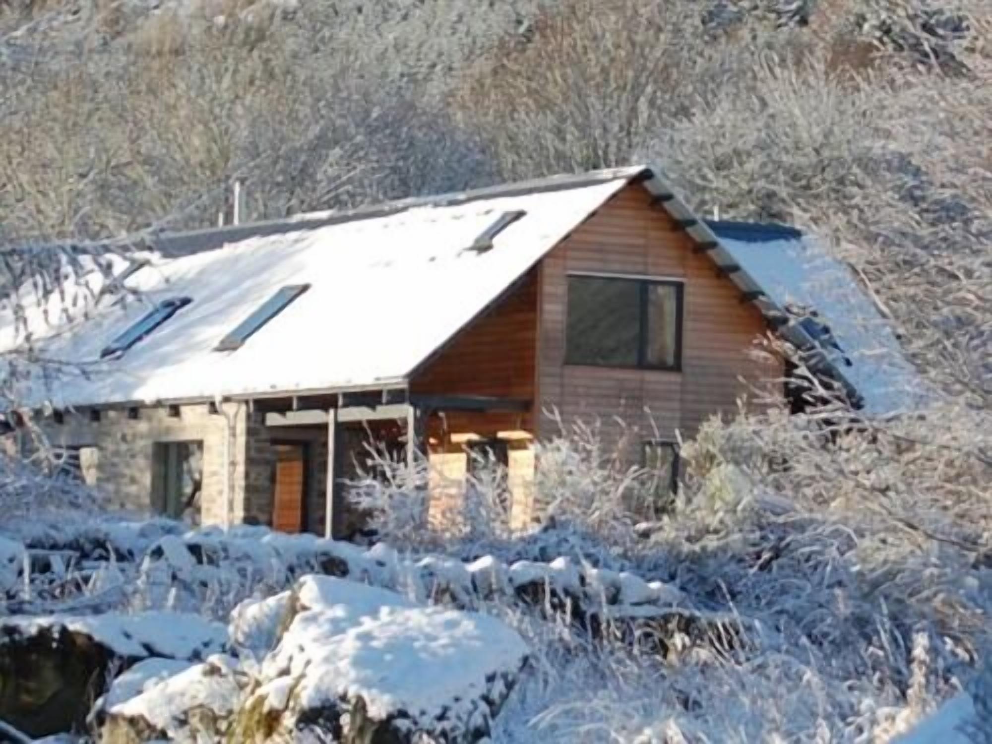 A wooden cabin with a sloped roof, surrounded by snow-covered ground and trees.