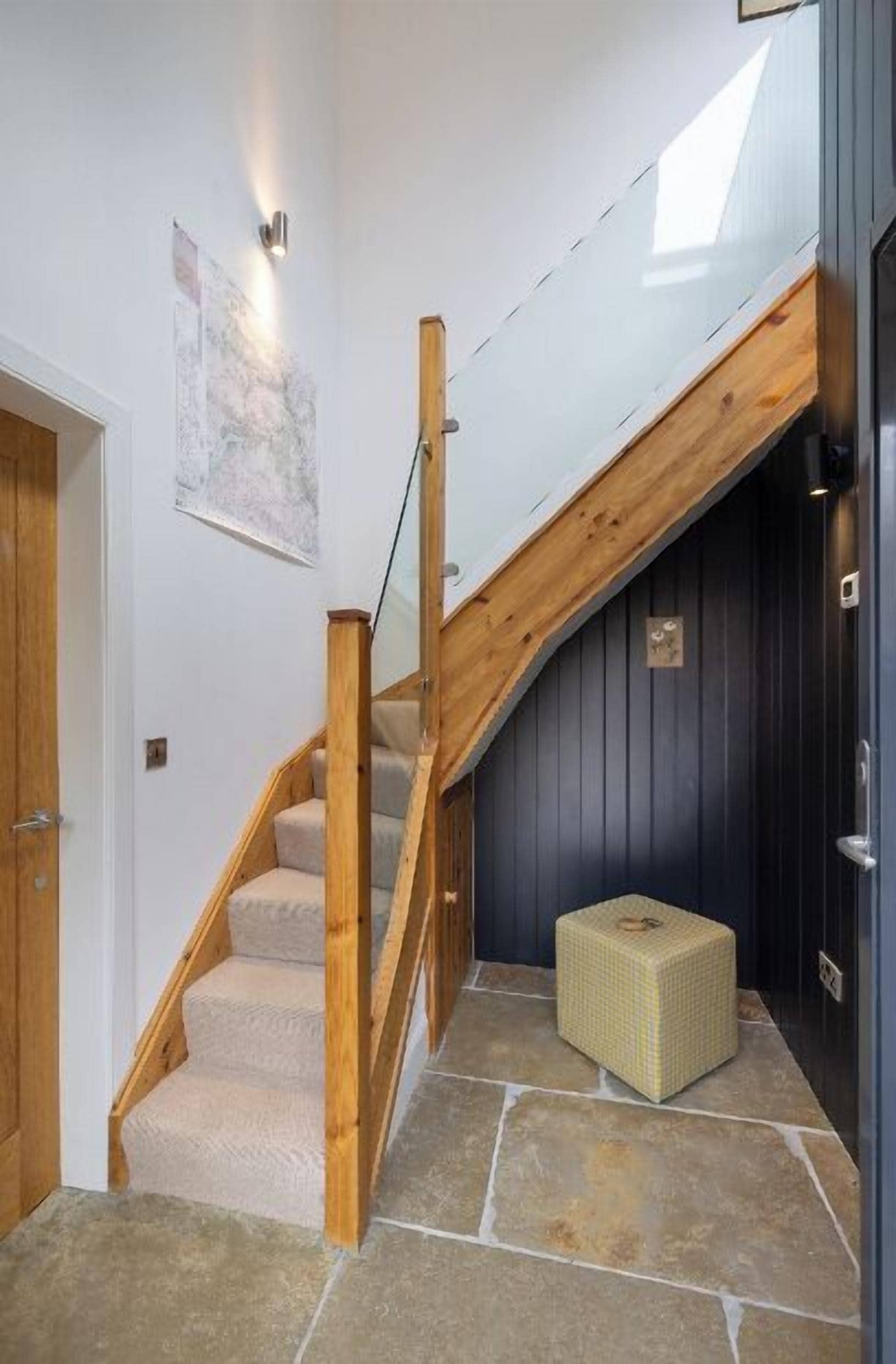 A staircase with wooden railings and beige carpeted steps leading to a dark room with a small yellow ottoman on a stone floor.