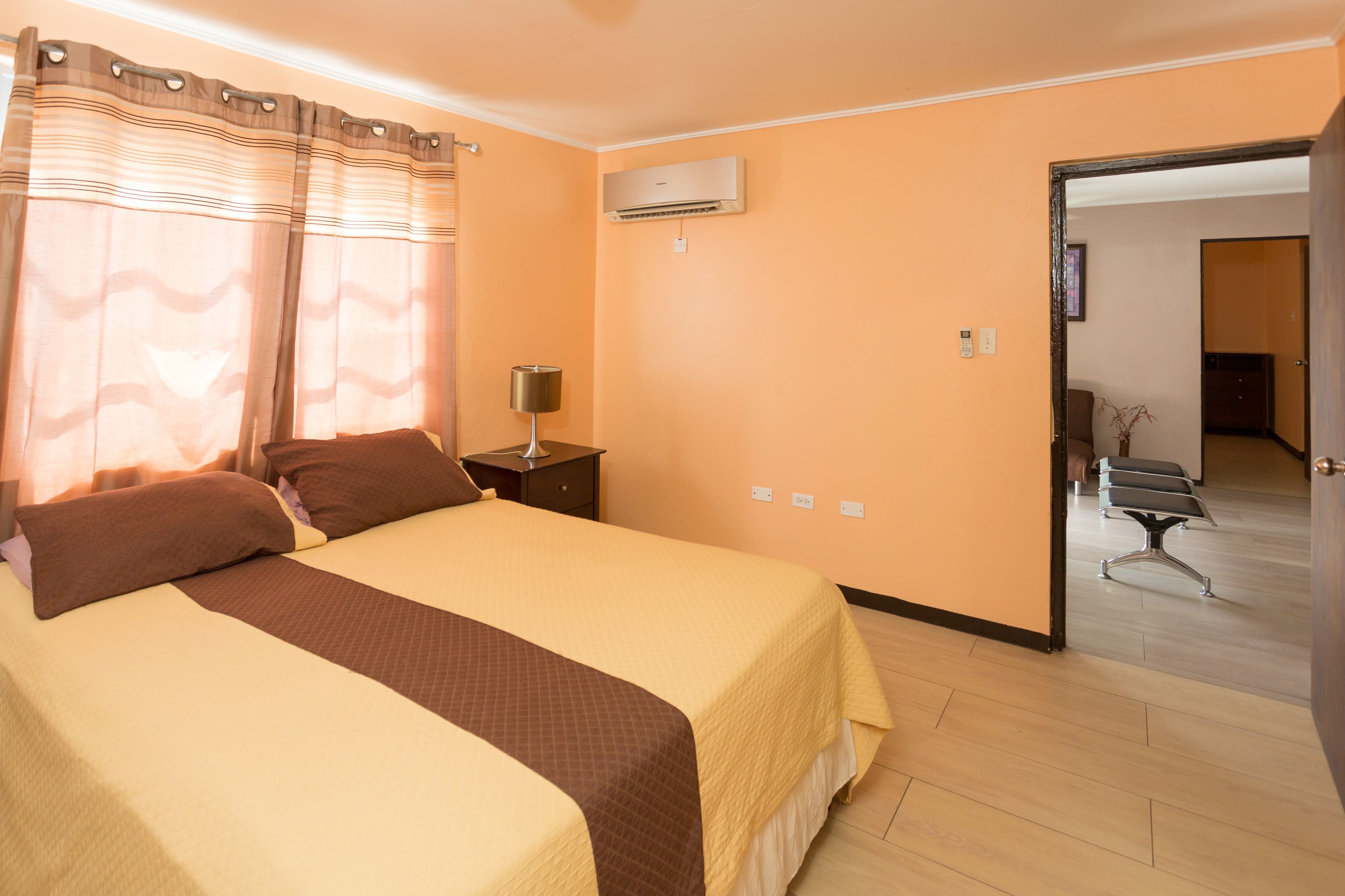 Een hotelkamer met een bed, een airconditioner, een nachtkastje en een deur die naar een andere kamer leidt.