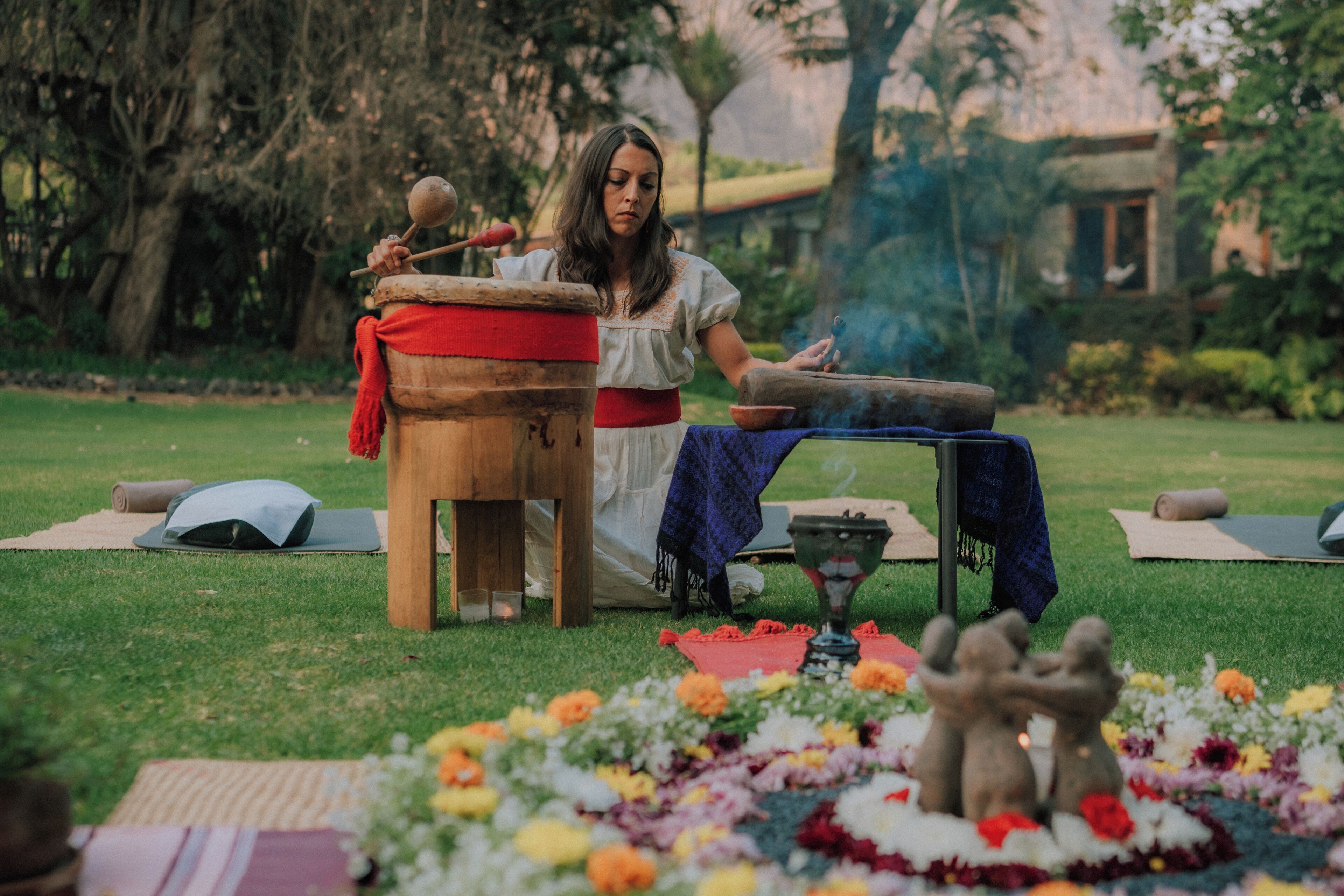 Una persona sentada al aire libre con un tambor, rodeada de tapetes de yoga, un cuenco y flores.