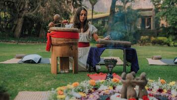 Una persona sentada al aire libre con un tambor, rodeada de tapetes de yoga, un cuenco y flores.