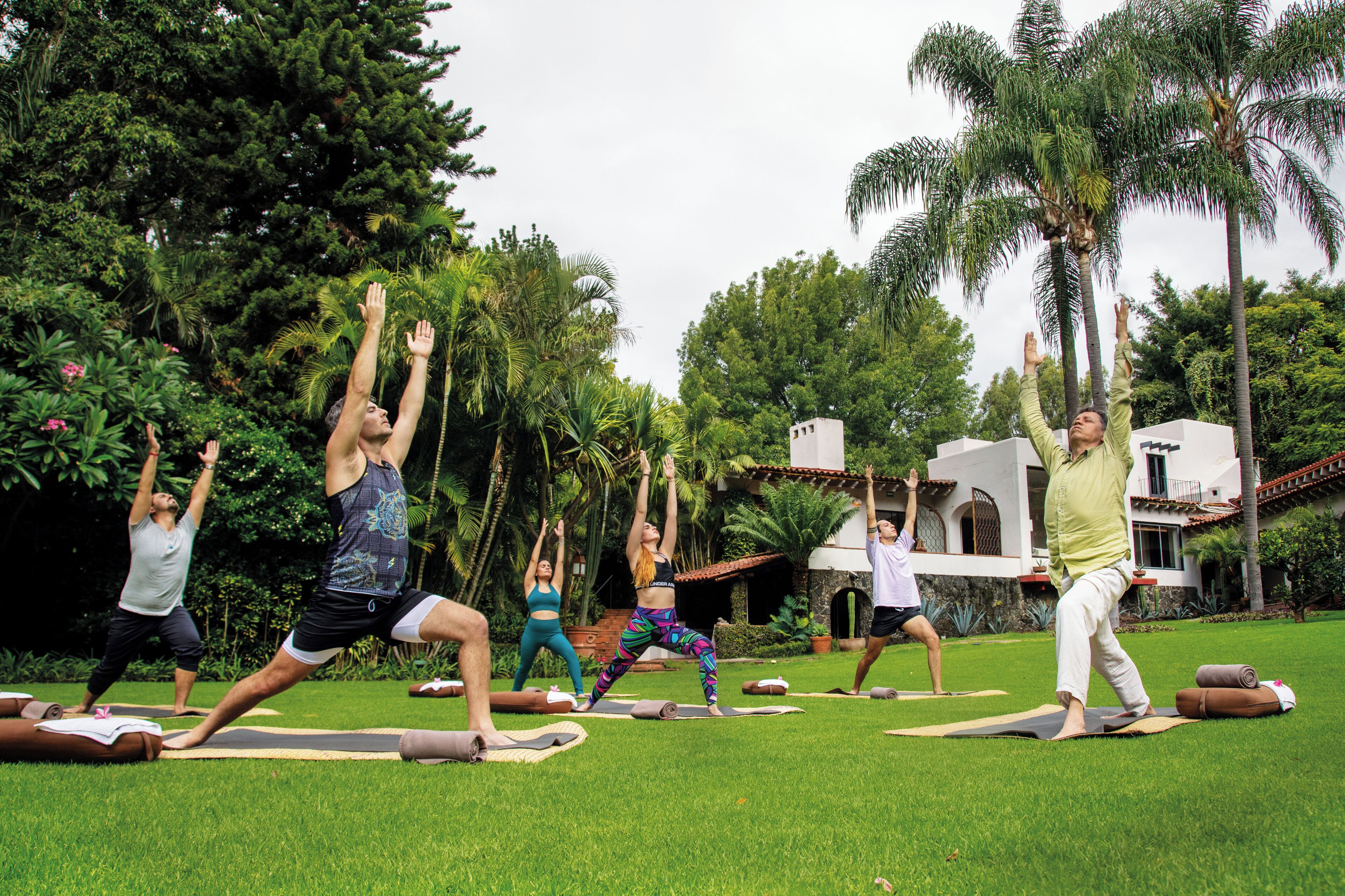 Un grupo de personas haciendo yoga sobre esterillas en un entorno al aire libre, con palmeras y un edificio al fondo.