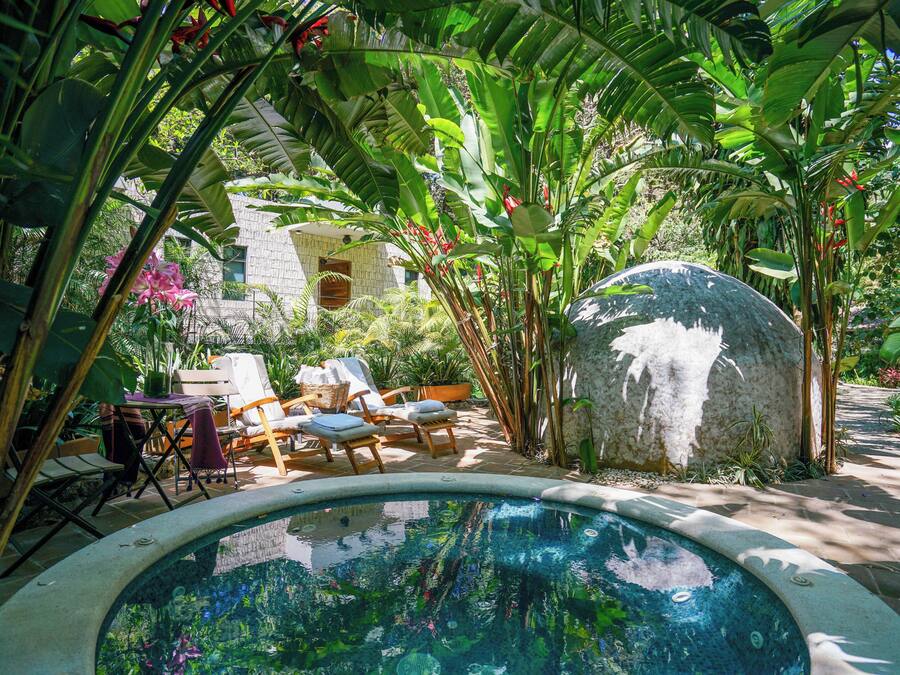 A pool surrounded by lush tropical plants, a stone structure, and a table with chairs.