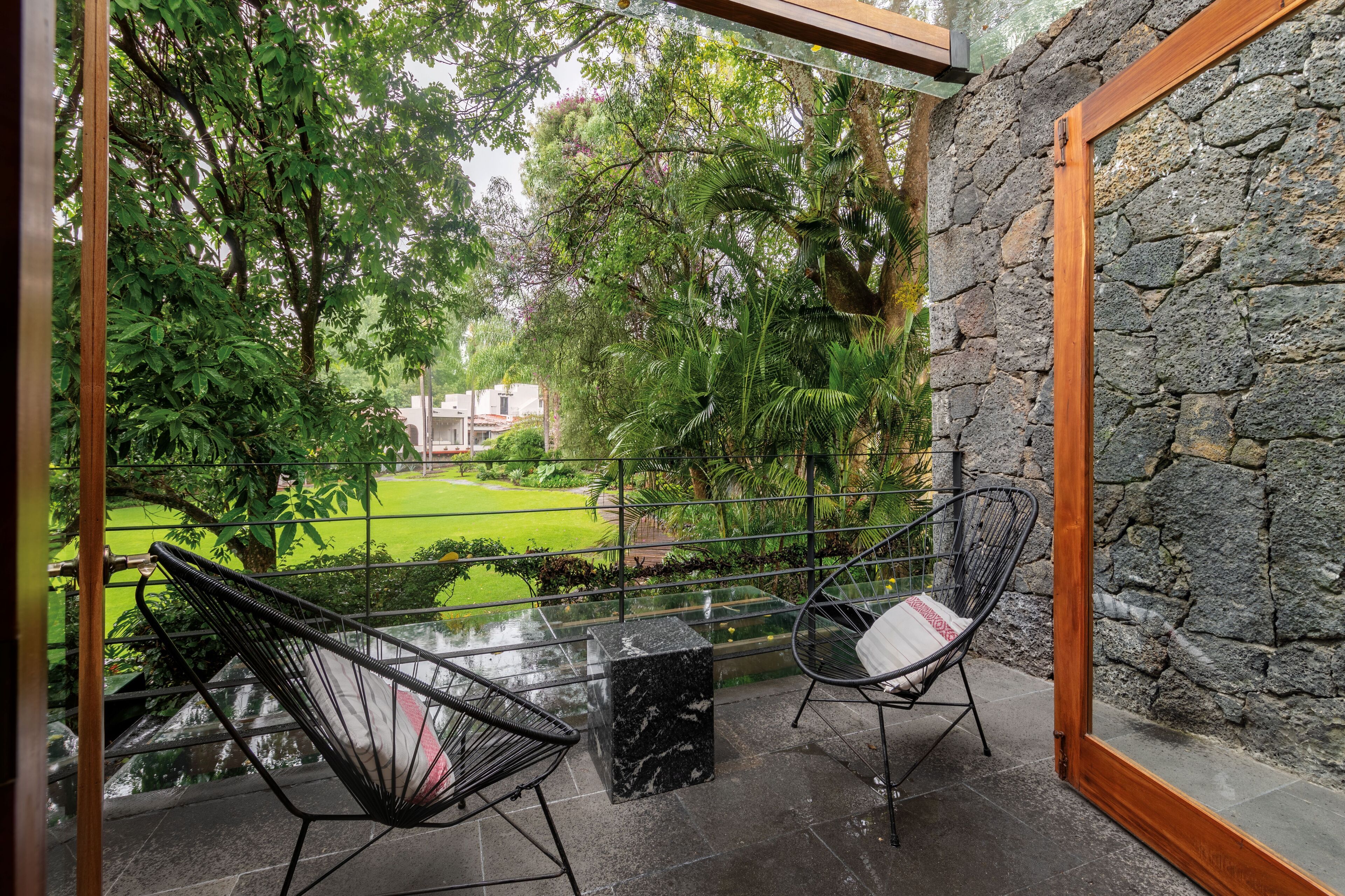 A balcony with two chairs, a marble table, and a view of a garden and buildings.