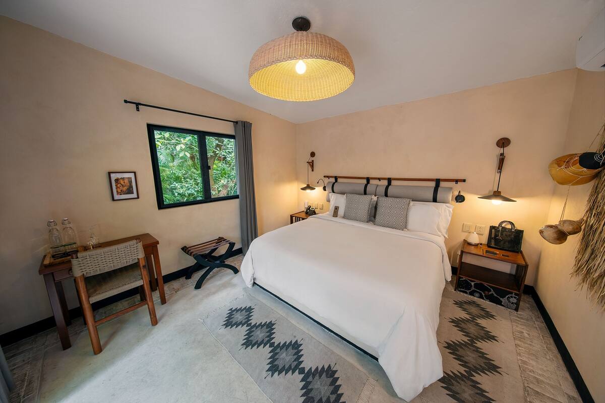 Une chambre avec un grand lit, des lampes de chevet, une fenêtre donnant sur un paysage verdoyant, une petite table avec une chaise et une décoration murale.