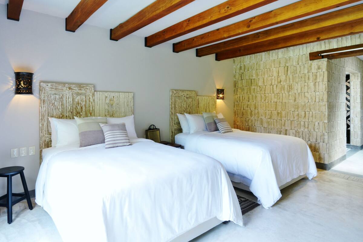 Une chambre d’hôtel avec deux lits, des poutres en bois au plafond, un mur en pierre et un petit tabouret.