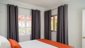 Quarto de hotel com cama, cortinas cinzas, uma janela e um cobre-leito laranja.