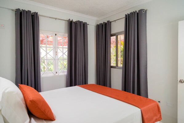 A hotel room with a bed, grey curtains, a window, and an orange bed runner.