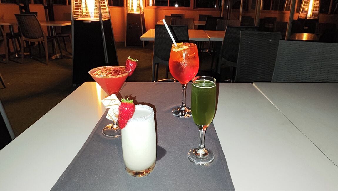 Ett bord med tre drinkar: en vit cocktail garnerad med en jordgubbe, en röd cocktail garnerad med en apelsinskiva och en grön cocktail med ett sugrör.