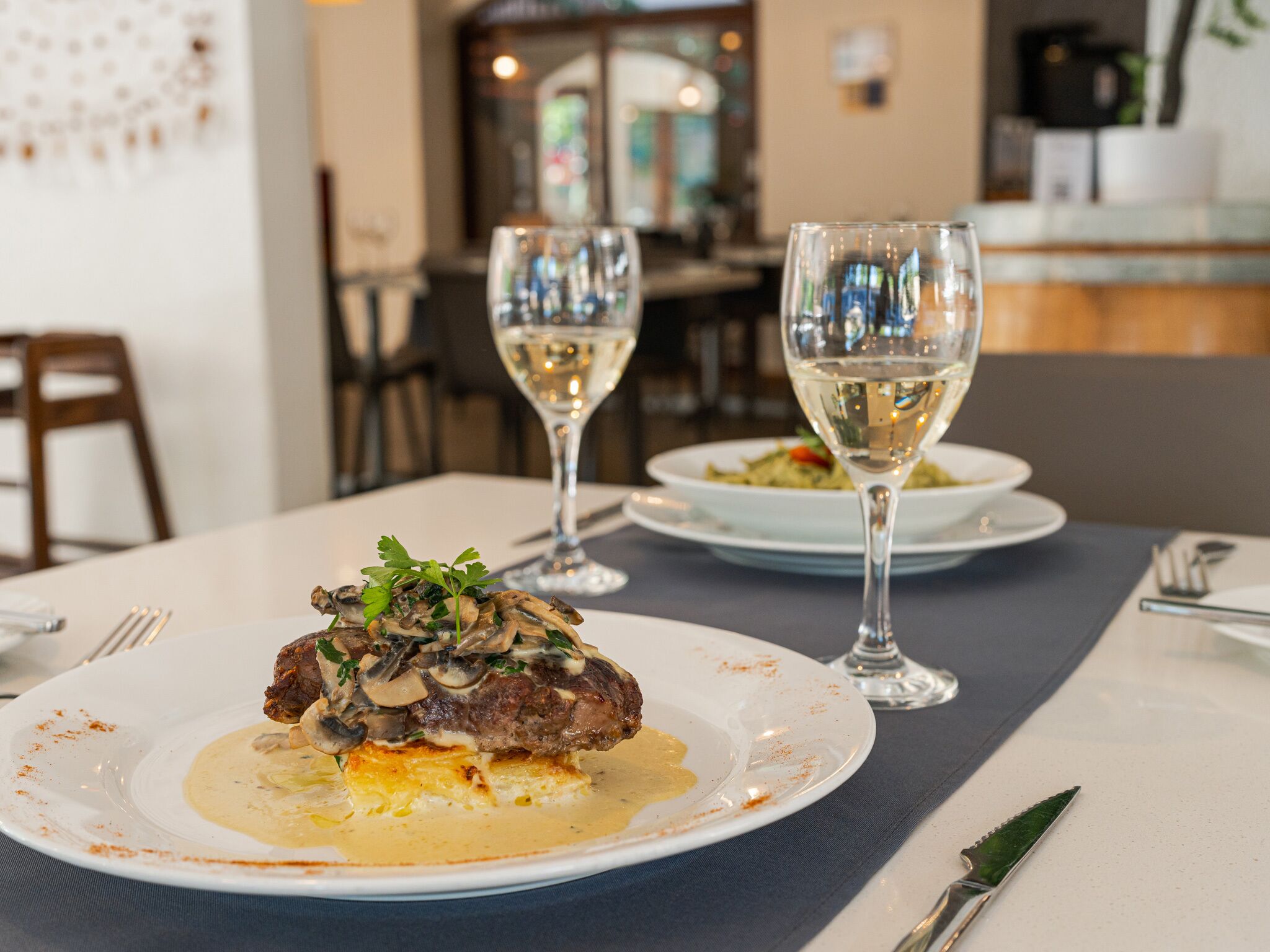 Une table à manger avec une assiette de steak garnie de champignons et nappée d’une sauce crémeuse, deux verres de vin blanc, ainsi qu’une fourchette et un couteau posés sur la table.