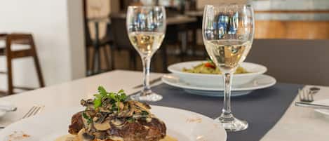 Une table à manger avec une assiette de steak garnie de champignons et nappée d’une sauce crémeuse, deux verres de vin blanc, ainsi qu’une fourchette et un couteau posés sur la table.
