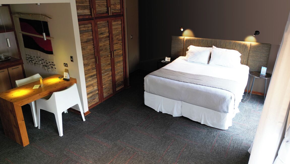 Quarto de hotel com cama, mesa de cabeceira, escrivaninha, cadeira e guarda-roupa.