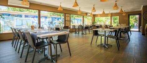 A spacious dining area with multiple tables and chairs, large windows offering views of greenery, and hanging pendant lights.