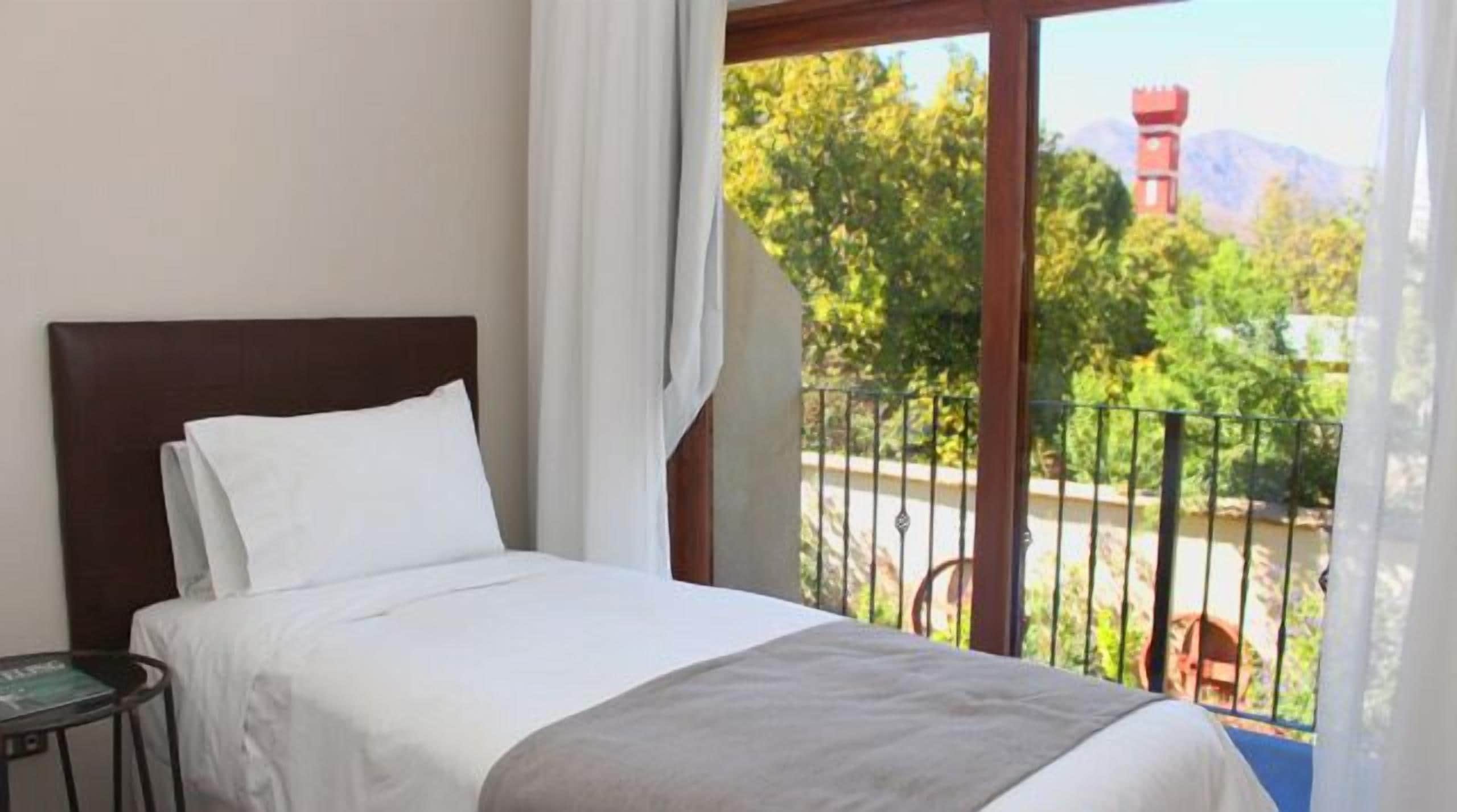 A hotel room with a bed, a small table, and a balcony with a view of trees and a building.