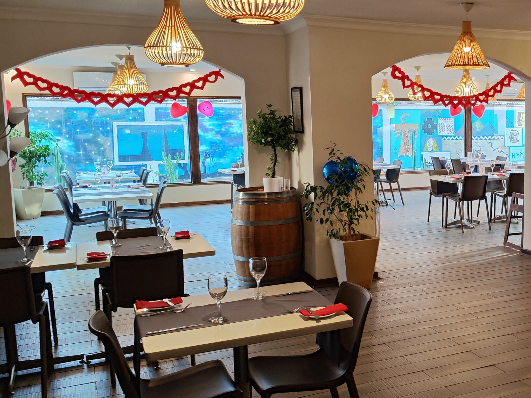 Un restaurante con mesas y sillas de madera, lámparas colgantes y vistas de la ciudad a través de amplios ventanales.
