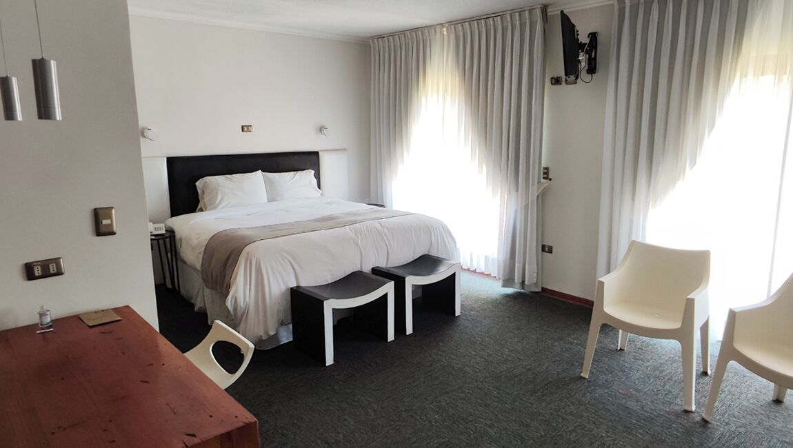 A hotel room with a bed, two white chairs, a wooden table, and a large window with curtains.