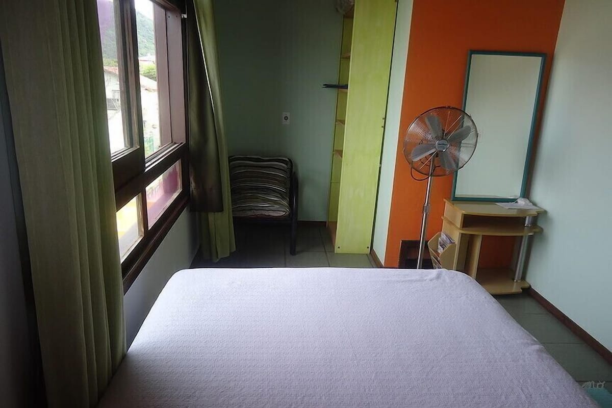 A room with a bed, a standing fan, a desk, and a chair.