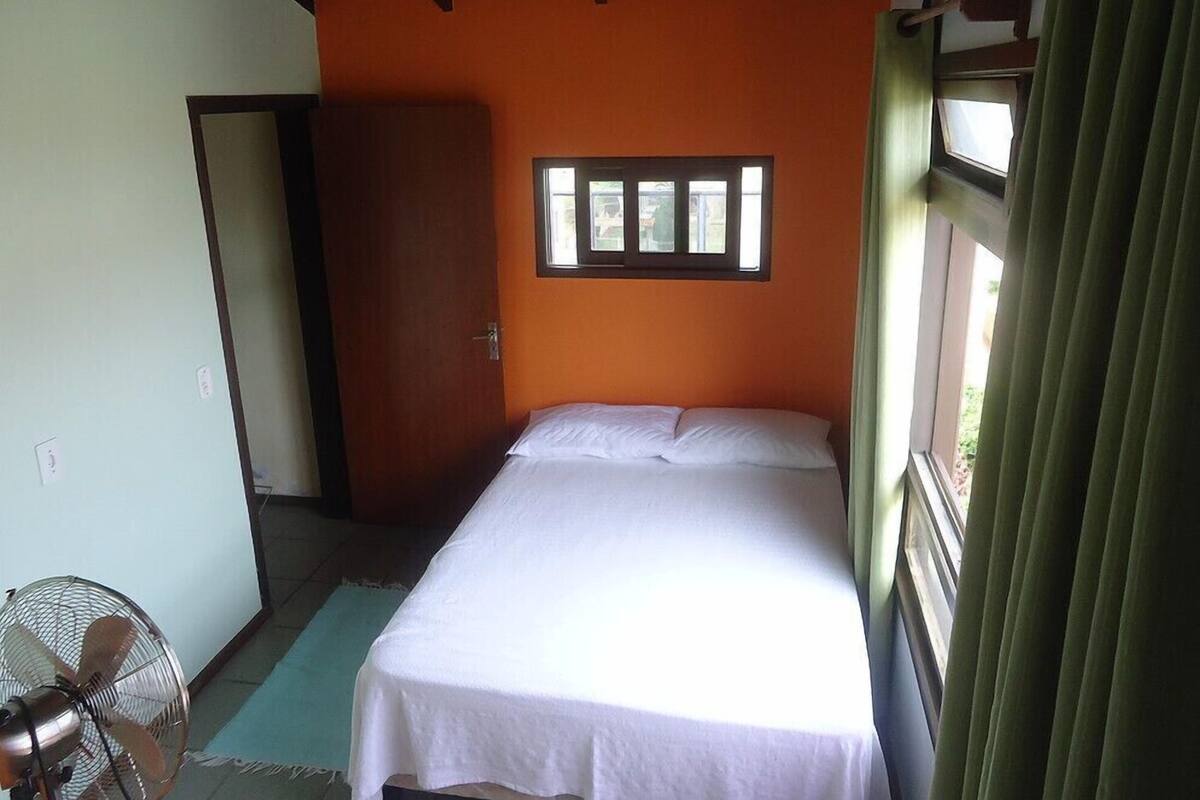 A single-bed room with an orange wall, a window with blinds, a standing fan, and a door to another room.