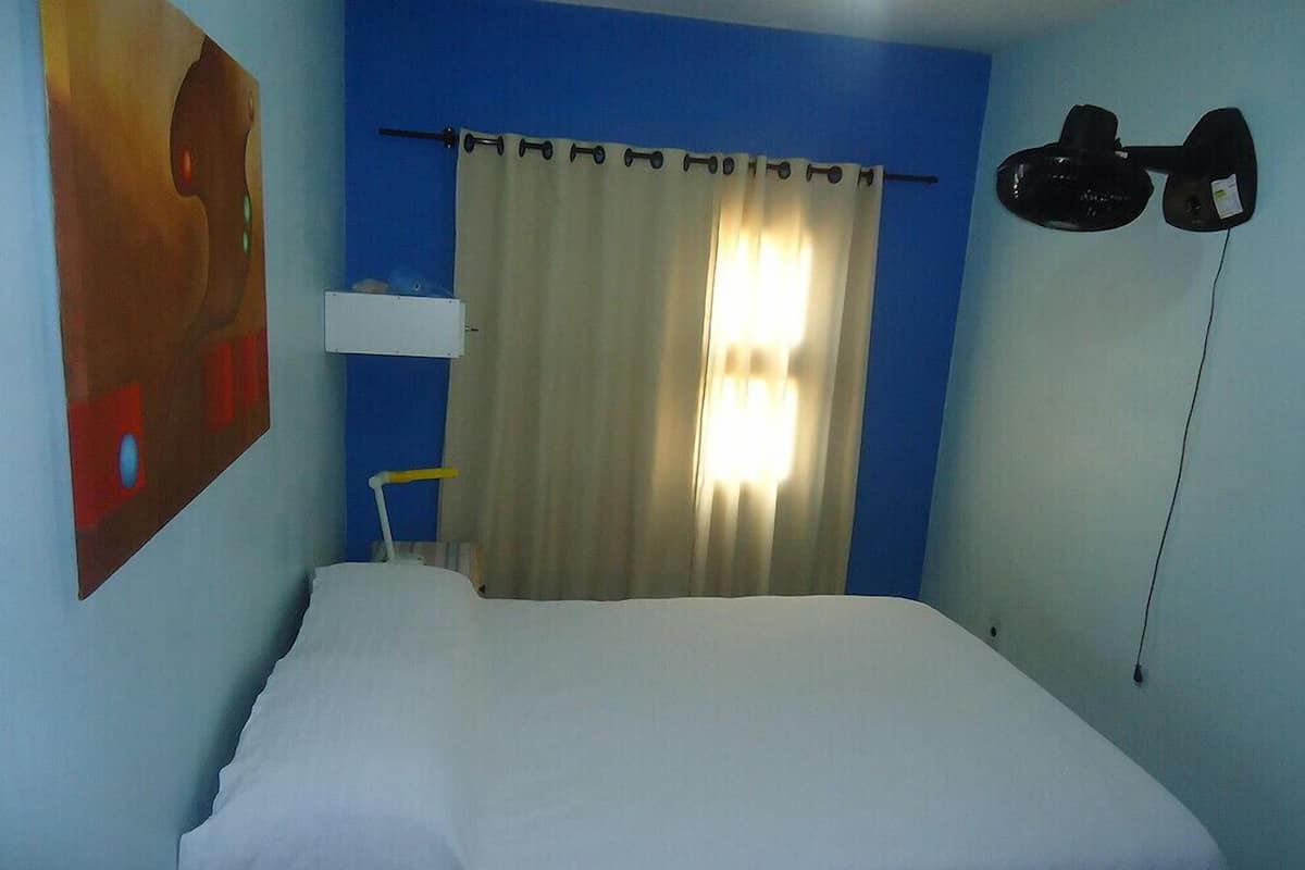 A hotel room with a bed, a curtain, a wall-mounted lamp, and a painting.